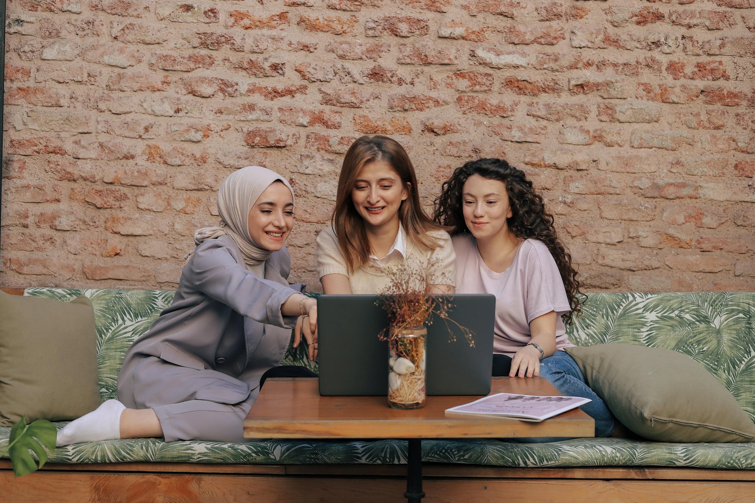 Three diverse women smiling, collaborating on a laptop in a cozy café with a rustic brick wall backdrop.